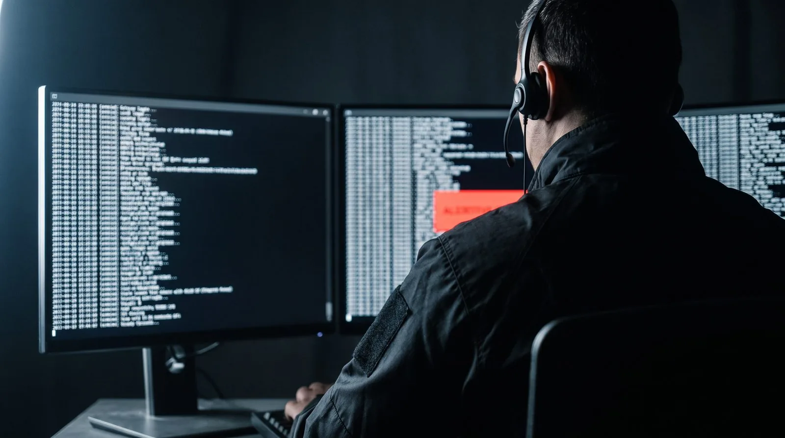 Security operator at three monitors of terminal output with one red alert panel